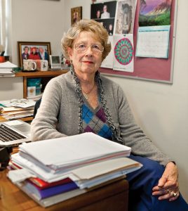 An elderly woman with glasses and short hair sits at a desk in a cluttered office. She is wearing a grey cardigan over a patterned top. The desk has a stack of papers and a laptop, hinting at her adaptability for the future. Behind her, a bulletin board displays various photos and a calendar.