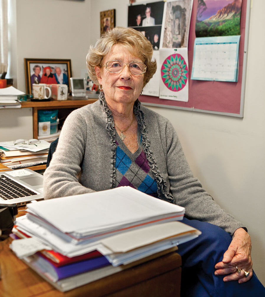 An elderly woman with curly hair and glasses sits in an office, surrounded by papers and a laptop on her desk. She's wearing a gray cardigan over a patterned sweater. Behind her are photos, a calendar, and colorful artwork pinned to the bulletin board.