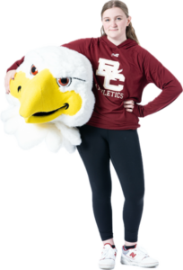 A person is standing with one hand on their hip, wearing a maroon BC Athletics hoodie and black leggings. They are holding a large eagle mascot head, which is characterized by a yellow beak and white feathers. Whether you're cheering with partners or parents, this look embodies team spirit.