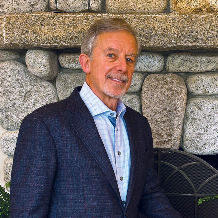 A man with short gray hair and a trimmed beard, wearing a dark blazer over a light blue checked shirt, stands in front of a stone fireplace, smiling slightly at the camera, ready to embrace the spirit of Fall 2025.