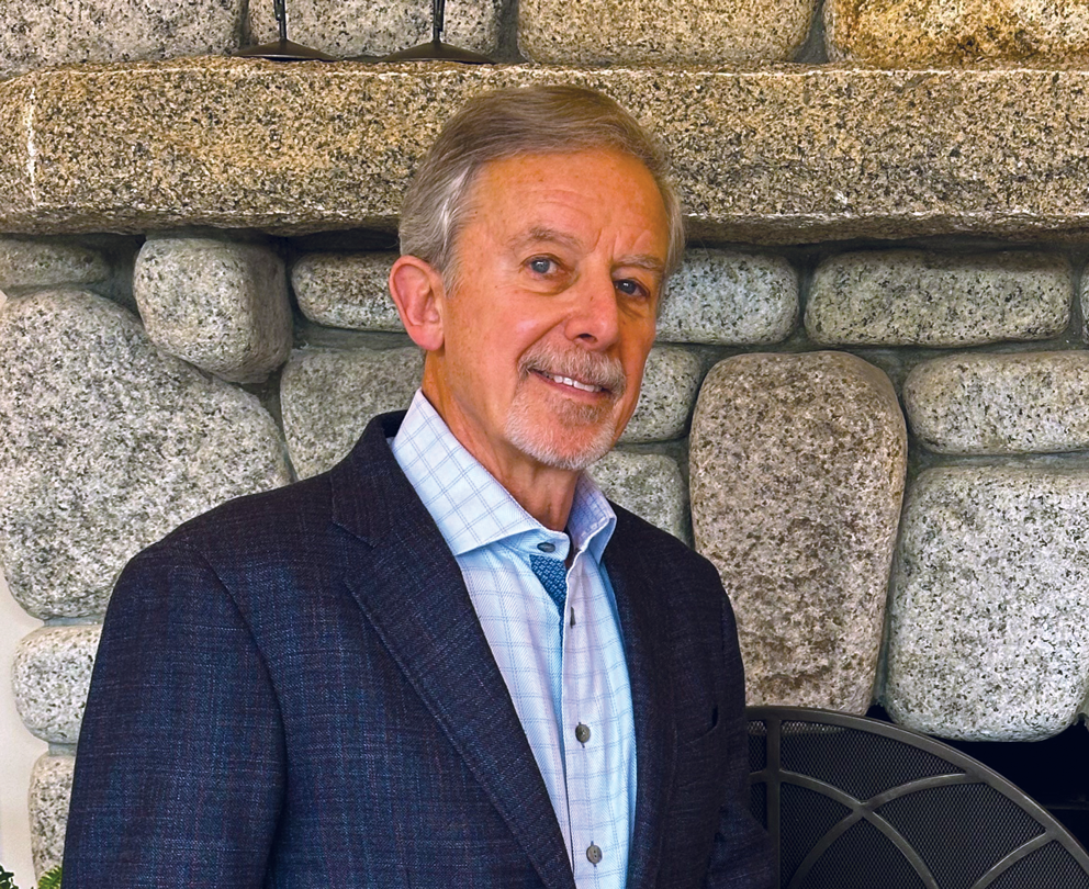 A man with short gray hair and a trimmed beard, wearing a checked shirt and dark blazer, stands in front of a stone fireplace, smiling slightly at the camera—an image of thinking like a lawyer and caring like an eagle.