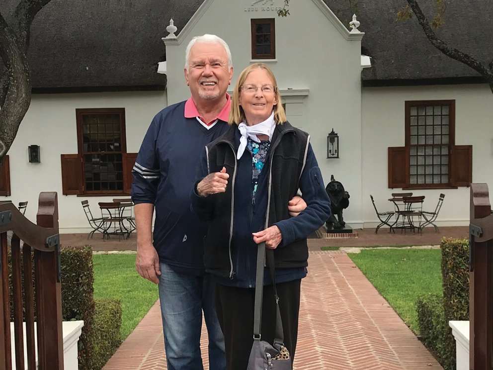 A smiling older man and woman stand arm-in-arm on a brick path in front of a white building with wooden windows and a thatched roof, with green lawns and outdoor tables visible on either side.