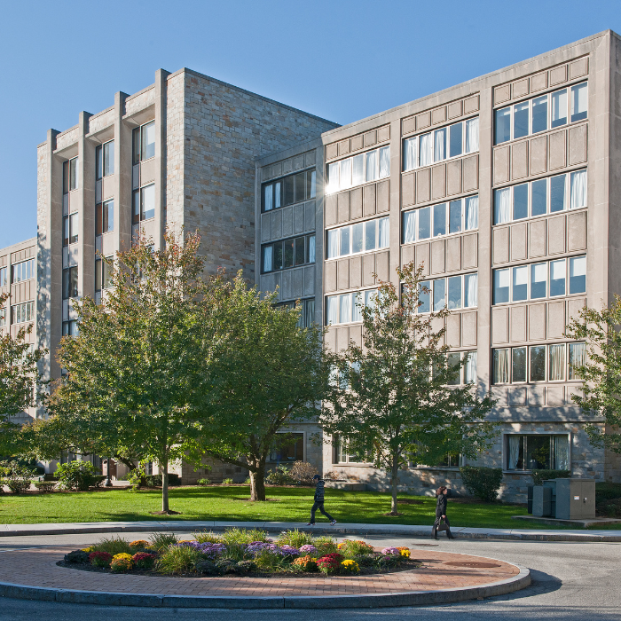 A large, multi-story academic building with many windows, trees, and a circular flower bed in front. Two people walk on the path near the building under a clear blue sky, hinting at the vibrant campus life of Fall 2025.
